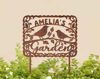 May include: A rusty metal garden sign with a decorative border and two birds perched on a branch. The sign reads "Amelia's Garden".