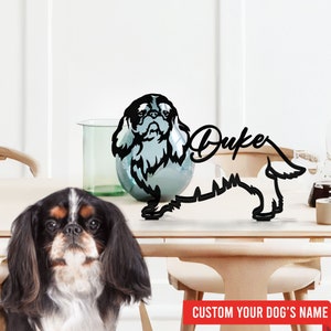 May include: Black metal silhouette of a Cavalier King Charles Spaniel dog with the name "Duke" written in cursive. The silhouette is on a wooden table with a real Cavalier King Charles Spaniel dog sitting in the foreground.