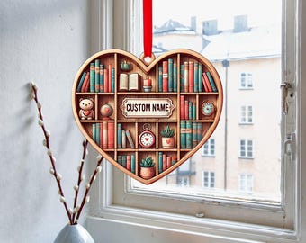 May include: A wooden heart-shaped ornament with a bookshelf design. The bookshelf is filled with books, a teddy bear, a clock, and a potted plant. The ornament has a red ribbon and a sign that says "CUSTOM NAME".