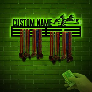 May include: A black metal medal display rack with a bobsled silhouette and the text "CUSTOM NAME". The rack has three horizontal bars for hanging medals. The rack is illuminated with green LED lights.