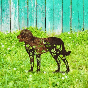 May include: A rusty metal silhouette of a dog with the name Max and the dates 2002-2023 on it. The silhouette is surrounded by green grass and a wooden fence.