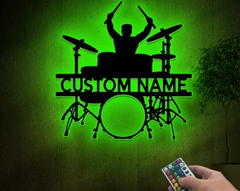 Pode incluir: Uma silhueta preta de um baterista tocando uma bateria com uma luz verde atrás. A bateria tem uma placa de identificação que diz "CUSTOMNAME". Um controle remoto está no canto inferior direito da imagem.