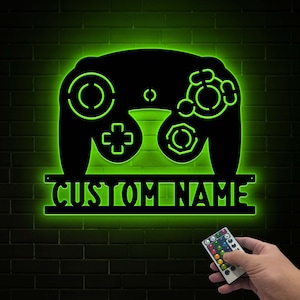 May include: A green neon light shaped like a video game controller with the text "CUSTOM NAME" underneath. The light is mounted on a brick wall. A remote control is visible in the bottom right corner of the image.