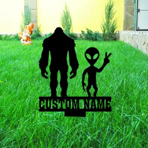May include: Black silhouette of a Bigfoot and an alien with a peace sign, standing on a green lawn with a sign that says "CUSTOM NAME".