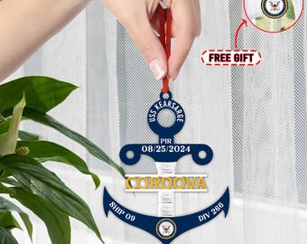 May include: A blue and white anchor-shaped Christmas ornament with the text "USS KEARSARGE" and "PIR 08/25/2024" on the top, "CORDOVA" in the middle, "SHIP 09" on the left, and "DIV 266" on the right. The ornament is hanging from a red ribbon.