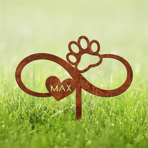 May include: A rusty brown metal garden stake featuring an infinity symbol, a heart with the name "MAX", and a paw print. The stake is designed to be placed in a garden or lawn, set against a backdrop of green grass.