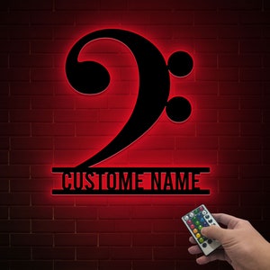 May include: Black metal bass clef wall art with red LED backlighting. The sign includes the text "CUSTOM NAME" and is mounted on a red brick wall. A remote control is visible in the image.