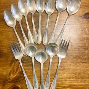 May include: A collection of vintage silverware, including spoons, forks, and ladles, arranged on a wooden surface. The silverware has a shiny, silver finish with decorative detailing on the handles. The spoons have an oval bowl shape, and the forks have four tines.