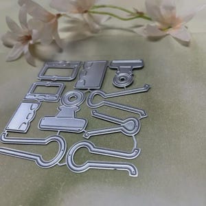 May include: A collection of silver metal die cuts in various shapes, including frames, a camera, and fasteners. The die cuts are arranged on a light green surface, with white flowers in the background. These are likely used for paper crafting or scrapbooking.