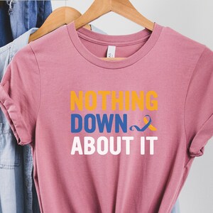 Op de afbeelding: Een roze T-shirt met de tekst "Nothing Down About It" in wit, blauw en geel. Een geel en blauw lint bevindt zich naast het woord "Down".