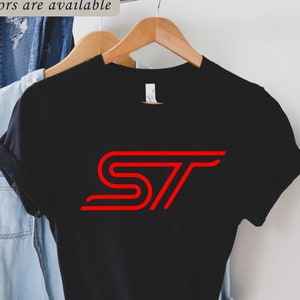 May include: Black t-shirt with a red "ST" logo design. The shirt is hanging on a wooden hanger. The text "Different colors are available" is at the top. The shirt is displayed with a denim shirt and a brown hat.