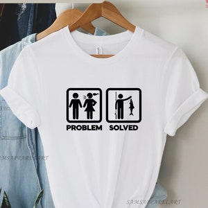 Può includere: T-shirt bianca con grafica nera raffigurante una coppia che litiga, con la scritta "PROBLEMA" e una persona che pesca, con la scritta "RISOLTO". Il testo "Sono disponibili diversi colori" è in alto. La maglietta è appesa a una gruccia di legno.