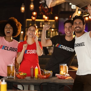 Puede incluir: Cuatro amigos con camisetas con los nombres "Clara", "Cora", "Carson" y "Daniel" impresos en ellas est&aacute;n brindando en un bar. Tienen bebidas y aperitivos.