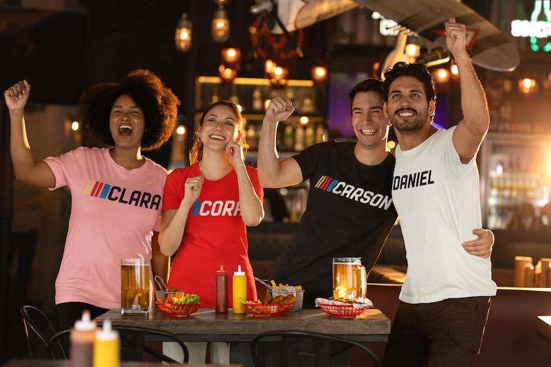 Puede incluir: Cuatro amigos con camisetas con los nombres "Clara", "Cora", "Carson" y "Daniel" impresos en ellas est&aacute;n brindando en un bar. Tienen bebidas y aperitivos.