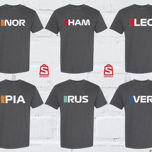 Puede incluir: Seis camisetas de color gris oscuro con texto blanco y detalles de color. Cada camiseta muestra una abreviatura de tres letras: NOR, HAM, LEC, PIA, RUS y VER. Las camisetas son de manga corta y de material suave.