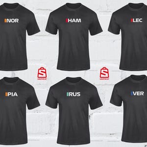 Puede incluir: Seis camisetas de color gris oscuro con diferentes textos de tres letras en varios colores. Los textos incluyen "NOR", "HAM", "LEC", "PIA", "RUS" y "VER". Cada camiseta tiene cuello redondo y mangas cortas.