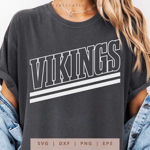 Może przedstawiać: Ciemnoszara koszulka z napisem "VIKINGS" w kolorze białym, obramowanym na czarno, oraz dwoma białymi poziomymi paskami. Koszulka ma swobodny styl, z podwiniętymi rękawami. Na dole obrazu widnieje napis "SVG | DXF | PNG | EPS".