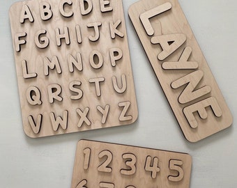 May include: Three light-colored wooden puzzle boards featuring cutouts. One board displays the alphabet from A to Z, another shows the numbers 0 through 9, and the third board features the name LAYNE in large, capital wooden letters.