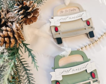 May include: Two green and white Christmas ornaments shaped like vintage cars with "Just Married" banners and red accents. The car on the bottom has the year 2024 printed on the back.