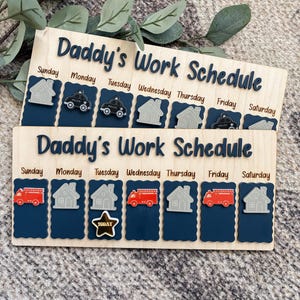 May include: Two rectangular wooden work schedule boards with the text Daddy's Work Schedule in dark blue. Each board features seven columns labeled Sunday through Saturday with dark blue scalloped sections and icons of houses, police cars, fire trucks, and a gold star labeled Today.