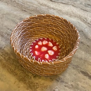 May include: A round, woven basket with a red and white interior. The basket is made of natural-colored fibers and has a circular design inside with a red background and white shapes.