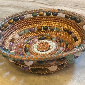 Può includere: Un cestino rotondo fatto a mano con un motivo a spirale. Il cestino è tessuto con fibre naturali e decorato con perline colorate nelle tonalità di verde, rosa, marrone e viola. Il bordo è di colore azzurro.