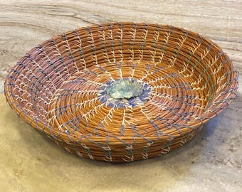 Op de afbeelding: Een ronde, ondiepe mand gemaakt van opgerolde dennennaalden in bruintinten, voorzien van decoratief stiksel in wit en blauw garen. Een kleine, getextureerde, lichtgroene keramische schijf is in het midden van de bodem geplaatst.