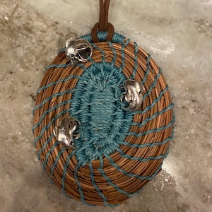 Könnte beinhalten: Handgefertigte Anhänger-Halskette mit einem gewebten ovalen Design aus braunen und türkisfarbenen Fasern. Der Anhänger ist mit drei silberfarbenen Charms und einer braunen Lederschnur versehen.