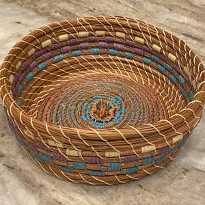 Puede incluir: Cesta redonda tejida a mano con un diseño en espiral. La cesta presenta un color marrón natural con acentos azules, morados y crema. El interior tiene un patrón en espiral con un diseño central en tonos azules y naranjas.