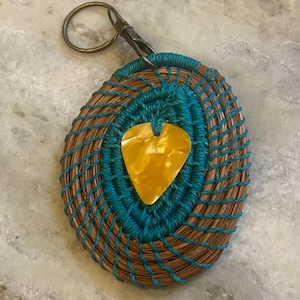 Könnte beinhalten: Handgefertigter ovaler Schlüsselanhänger mit einem gewebten braunen und türkisfarbenen Design. Ein gelber herzförmiger Anhänger befindet sich in der Mitte. Der Schlüsselanhänger hat einen Metallverschluss und einen Ring.
