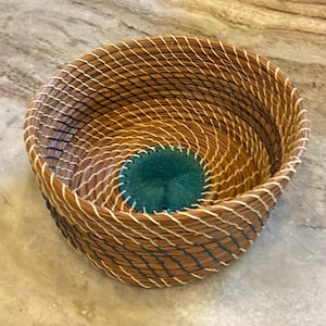 May include: A round, handwoven basket with a natural brown and cream color scheme. The basket features a teal-colored, circular accent in the center and is constructed with a diagonal weave pattern.