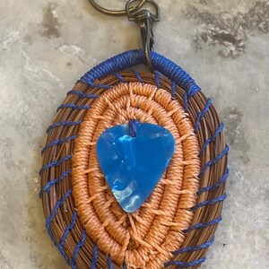 Könnte beinhalten: Handgefertigter ovaler Schlüsselanhänger mit einem gewebten Design in Braun- und Orangetönen, akzentuiert mit blauem Faden. Ein blauer Gitarrenplektrum ist mittig angebracht, mit Metallverschluss und Ring.