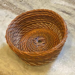 Pode incluir: Uma cesta redonda tecida à mão feita de agulhas de pinheiro secas. O recipiente em forma de tigela apresenta um padrão em espiral de agulhas castanhas e bronzeadas unidas por um fio fino de cor clara, criando uma superfície tecida texturizada.