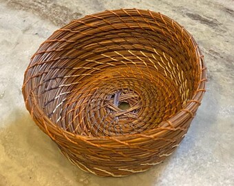 May include: A round, hand-coiled basket constructed from dried pine needles. The bowl-shaped vessel features a spiralling pattern of brown and tan needles bound together with thin, light-coloured thread, creating a textured, woven surface.
