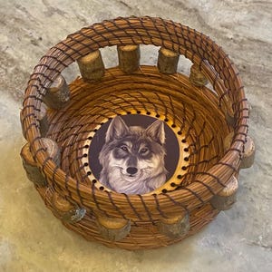 Könnte beinhalten: Ein handgefertigter, runder Korb aus Naturfasern. Der Korb hat ein detailliertes Wolfsporträt am Boden und ist mit kleinen Holzstücken am Rand verziert. Der Korb hat eine warme braune Farbe.
