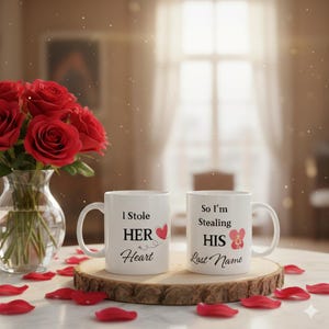 May include: Two white ceramic mugs with black text and red heart graphics. One mug reads "I Stole HER Heart," and the other says "So I'm Stealing HIS Last Name." The mugs sit on a wooden coaster, surrounded by red rose petals.