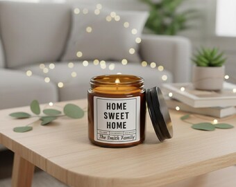 May include: A lit, brown glass jar candle with a black lid, sitting on a wooden table. The candle has a white label with the words "HOME SWEET HOME" and "The Smith Family" printed on it. The background includes a couch and a small plant.