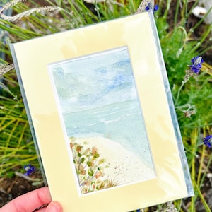 May include: A watercolor painting of a beach scene, framed in a pale yellow mat. The artwork depicts a sandy shore, turquoise water, and a blue sky. The painting is protected by a clear plastic sleeve, held against a background of green foliage.