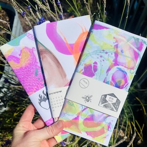 May include: Three colorful notebooks with abstract designs in pink, purple, and yellow. Each notebook is wrapped with a white band featuring a bee and the words "FOR YOU". One notebook has a calendar and a flame graphic.