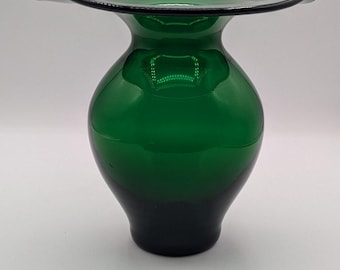 Puede incluir: Un jarrón de vidrio verde con un borde acampanado y un cuerpo redondeado. El jarrón tiene un color verde oscuro que se desvanece a un tono más profundo en la base. El borde está delineado con un fino borde negro.
