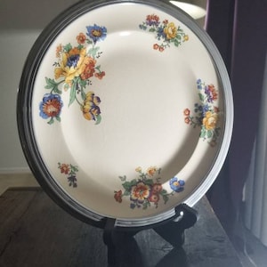 May include: A white ceramic plate with a silver rim and a floral design in shades of blue, yellow, orange, and green. The plate is on a black wooden stand.