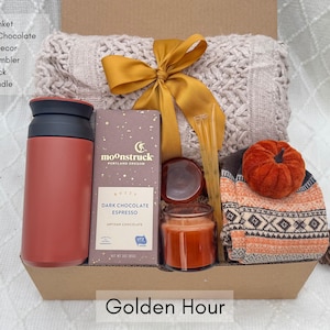 May include: A gift box with a throw blanket, gourmet chocolate, pumpkin decor, a coffee tumbler, a Nordic sock, and a scent candle. The box is tied with a gold ribbon and includes a candle and a chocolate bar. The text "Golden Hour" is at the bottom.