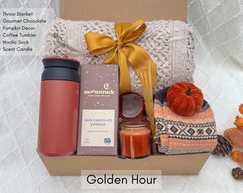 May include: A gift box with a throw blanket, gourmet chocolate, pumpkin decor, a coffee tumbler, a Nordic sock, and a scent candle. The box is tied with a gold ribbon and includes a candle and a chocolate bar. The text "Golden Hour" is at the bottom.