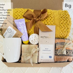 May include: A gift basket filled with self-care items, including a yellow throw blanket, a white coffee mug, a bar of soap, a chocolate bar, a bath bomb, and a tube of lavender sage hand cream. The basket is labeled "Big Hug".
