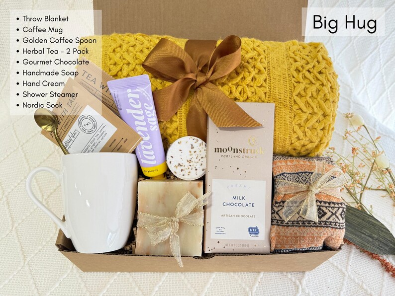 May include: A gift basket filled with self-care items, including a yellow throw blanket, a white coffee mug, a bar of soap, a chocolate bar, a bath bomb, and a tube of lavender sage hand cream. The basket is labeled "Big Hug".