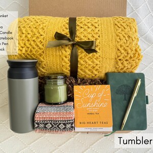 K&ouml;nnte beinhalten: Eine Geschenkbox mit einer gelben Decke, einem grauen Becher, Kr&auml;utertee, einer Duftkerze, einem Notizbuch, einem goldenen Stift und einer nordischen Socke. Die Box ist mit "Tumbler Box" beschriftet.