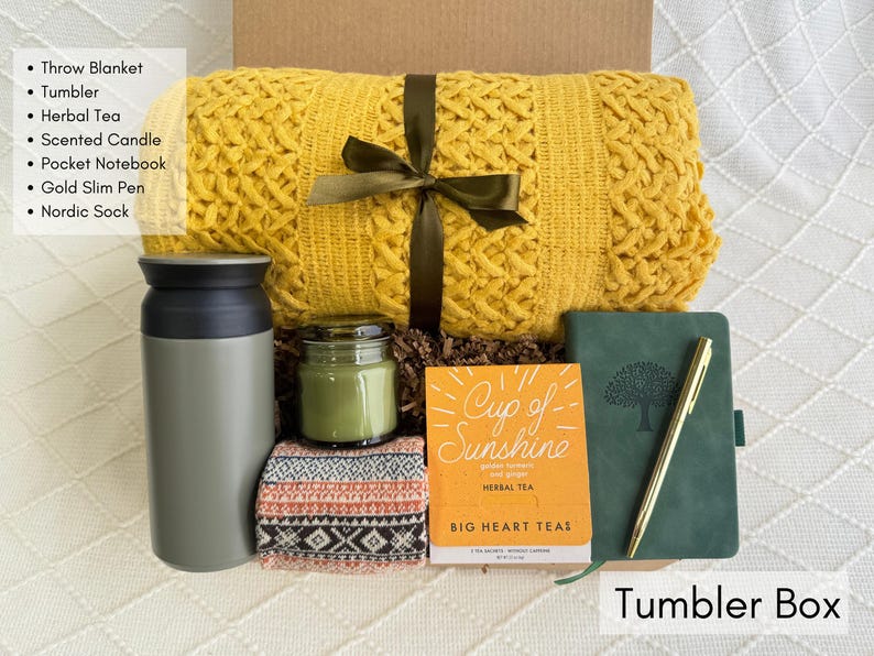 K&ouml;nnte beinhalten: Eine Geschenkbox mit einer gelben Decke, einem grauen Becher, Kr&auml;utertee, einer Duftkerze, einem Notizbuch, einem goldenen Stift und einer nordischen Socke. Die Box ist mit "Tumbler Box" beschriftet.