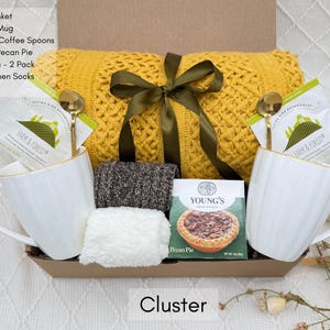 May include: A cozy gift basket filled with a yellow throw blanket, two white mugs with gold trim, two gold spoons, a pecan pie, a package of herbal tea, and socks. The basket is tied with a green ribbon and the words "FARM & FOREST" and "YOUNG'S" are visible on the packaging.