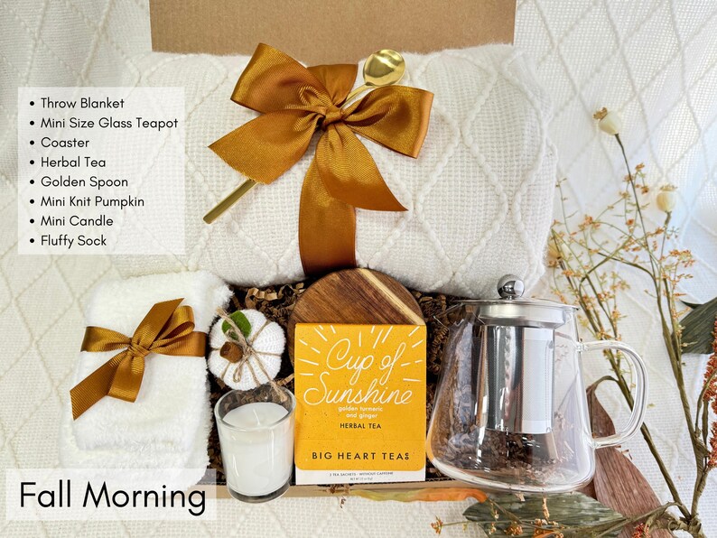 K&ouml;nnte beinhalten: Eine Geschenkbox mit einer wei&szlig;en Decke, einer Mini-Glas-Teekanne, Kr&auml;utertee und anderen Artikeln. Die Box enth&auml;lt einen goldenen L&ouml;ffel, einen Mini-Strickk&uuml;rbis, eine Kerze und flauschige Socken. Der Text "Fall Morning" und "Cup of Sunshine" sind sichtbar.