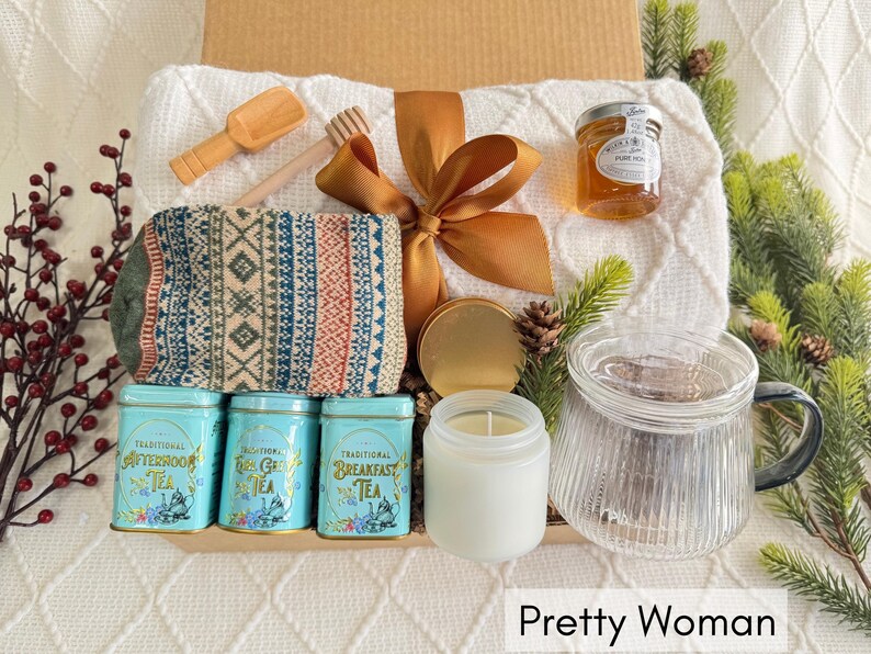 May include: A gift basket filled with a variety of items, including a white blanket, a pair of knitted socks, a jar of honey, a candle, a tea infuser, and three boxes of tea. The basket is decorated with a brown ribbon and a gold bow.